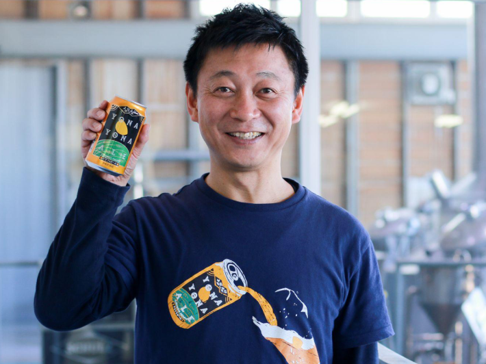 Naoyuki Ide (Tencho), President and CEO, smiling and holding a can of Yona Yona Ale with the brewery in the background